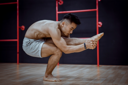 a muscular asian man without a shirt but with grey shorts on holding a pistol squat and holding his free leg with his hands