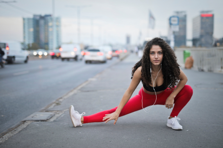 fit healthy latin woman with curly brown hair in red leggings and black sports bra with headphones in her ears wearing white running shoes stretching her legs after a good run