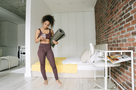 fit healthy black woman wearing brown leggings and brown sports bra checking her phone for her exercise routine while holding her yoga mat in her studio apartment