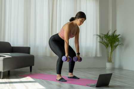 fit healthy asian woman in black leggings and pink tank top doing deadlifts with purple dumbbells on her pink yoga mat in front of her lap top at home