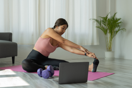 fit asian woman wearing black leggings and pink tank top stretching her legs on a pink yoga mat in front of her laptop and a purple set of dumbbells at her home