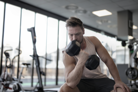 muscular white man with white cut-off t-shirt and black shorts doing seated dumbbell bicep curls at a gym