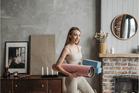 fit healthy woman wearing beige leggings and tank top holding a yoga mat and blocks at her home