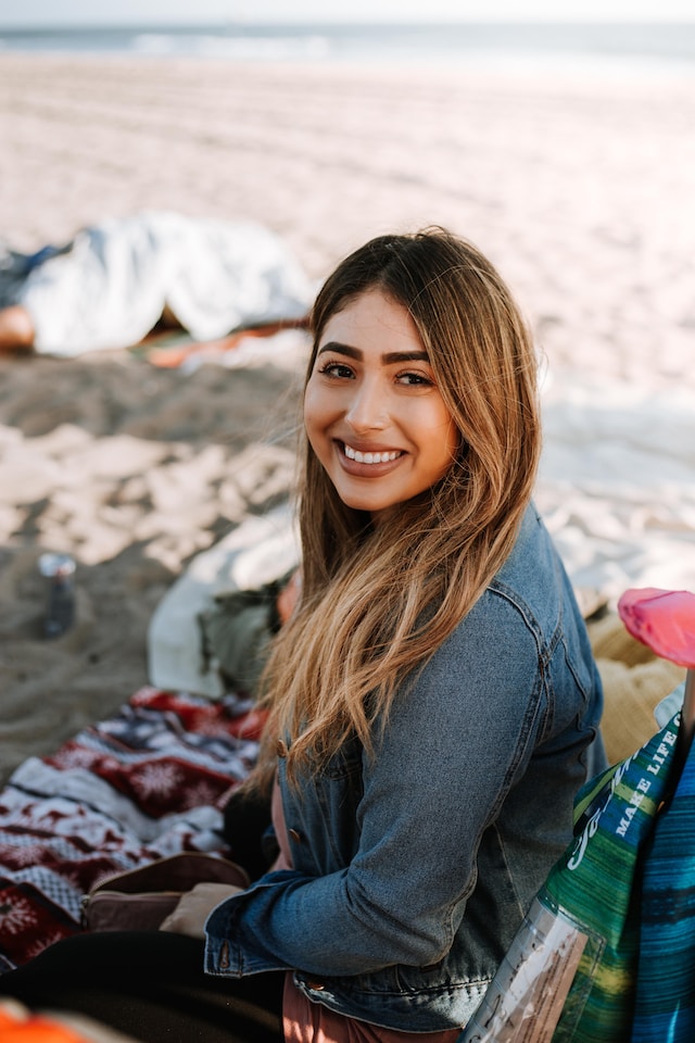 woman with brown hair and jean jack sitting on the beach smiling