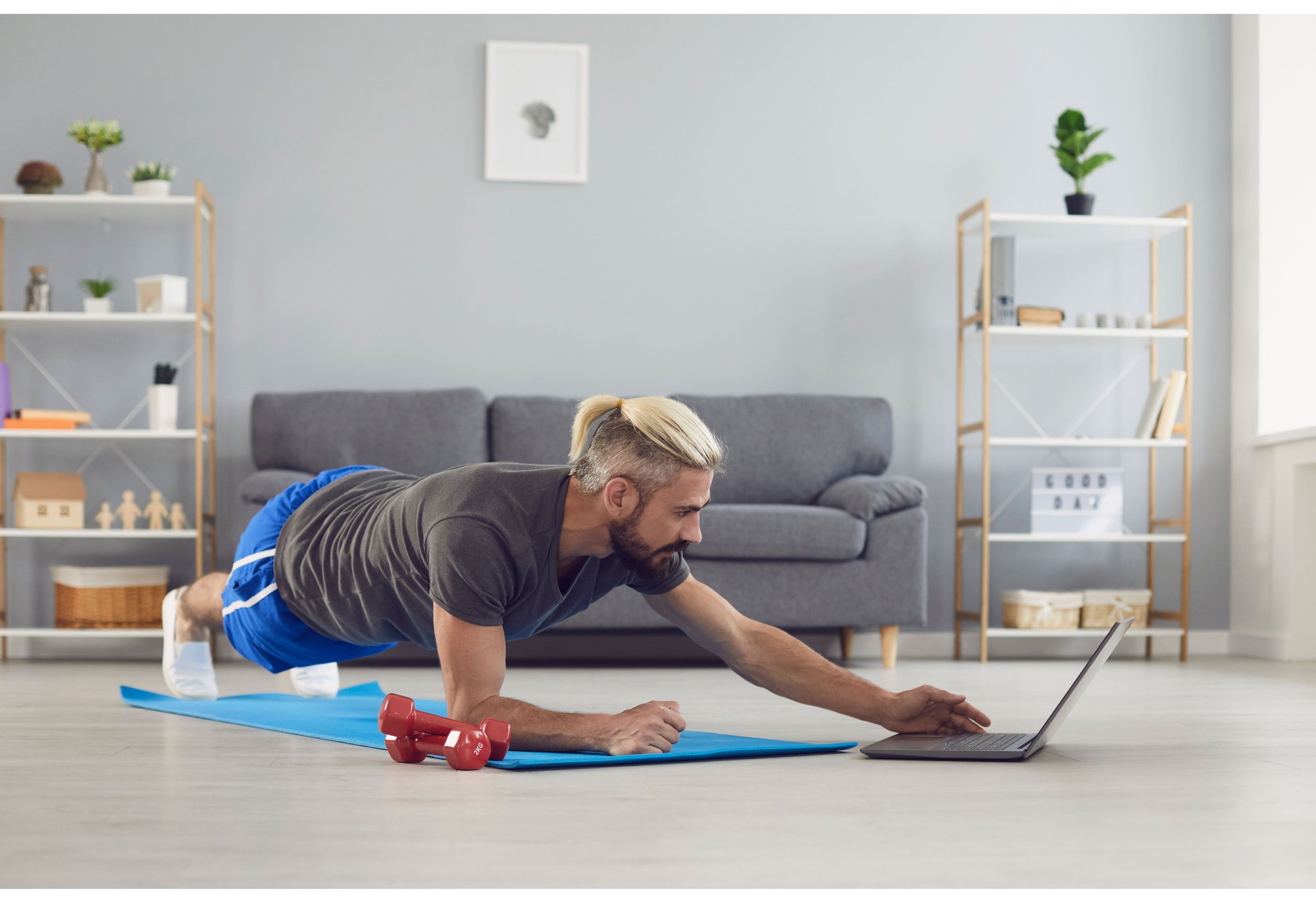 fit healthy man with blue shorts and grey t-shirt doing a plank on his yoga mat in front of his laptop at home