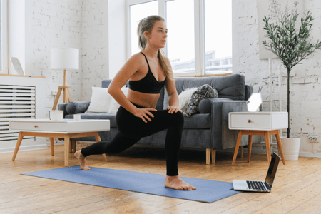 blonde woman wearing black leggings and black sports bra holding a lunge on her blue yoga mat in front of her laptop in her home