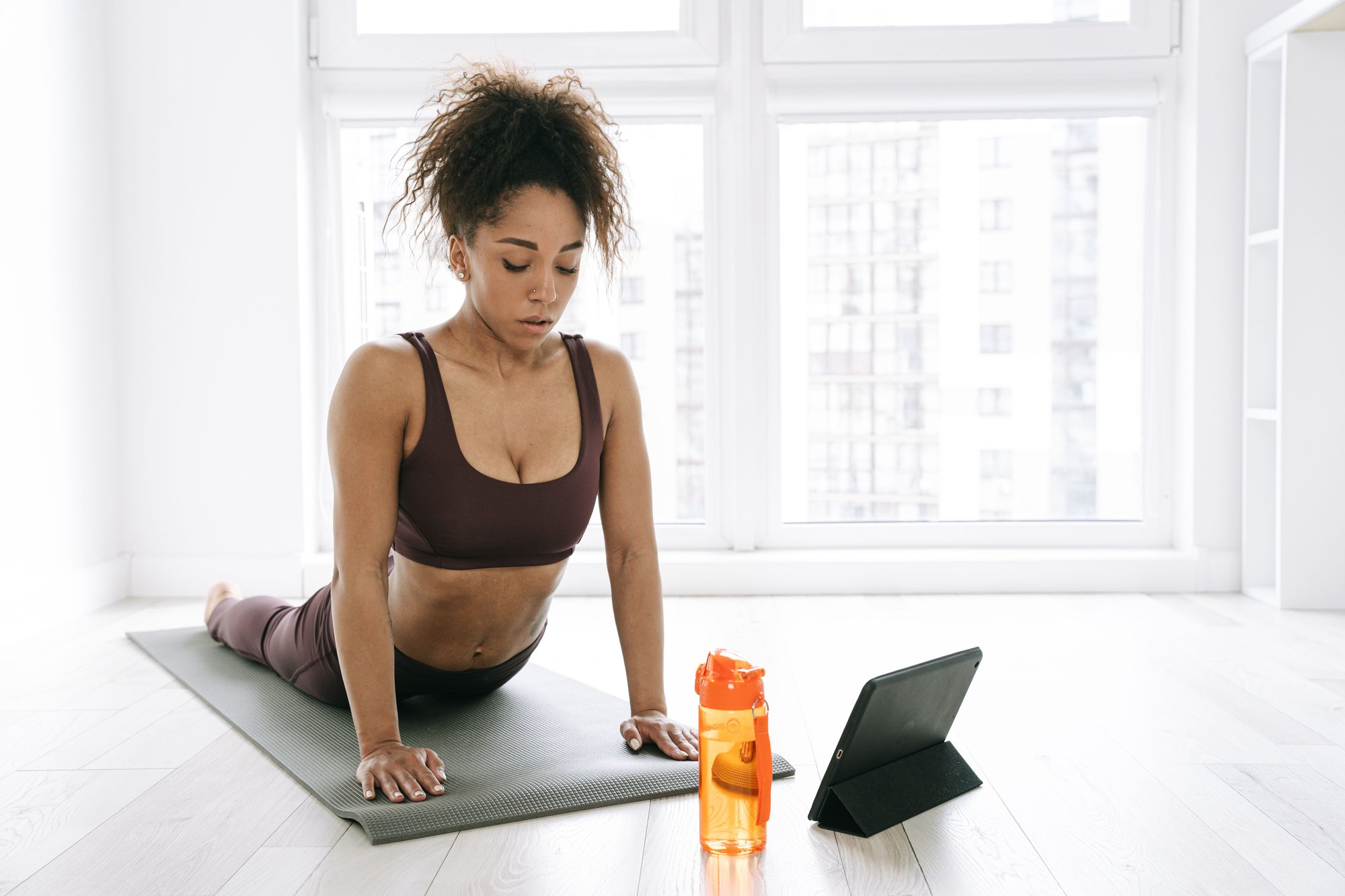 fit latin woman in brown leggings and brown sports bra stretching on a yoga mat following her personal trainer on her tablet