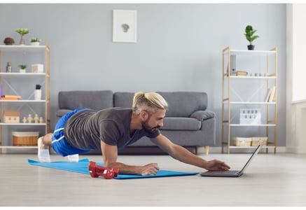 man with a blonde man bun wearing a grey t-shirt and blue shorts holding a plank on a blue yoga mat with red dumbbells on the side in check his laptop at his home