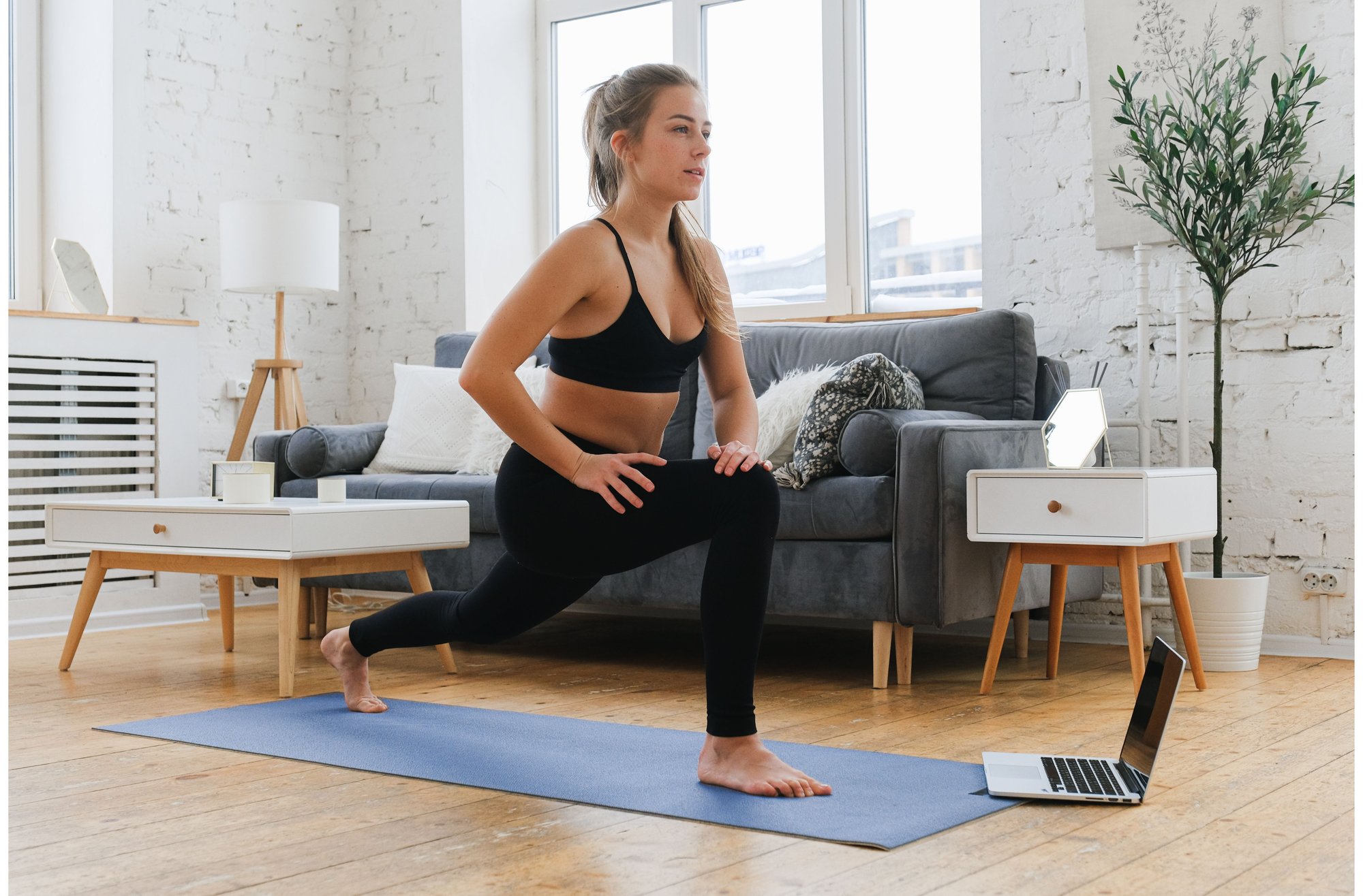 blonde woman wearing black leggings and black sports bra doing lunges on her blue yoga mat in front of her laptop at her home