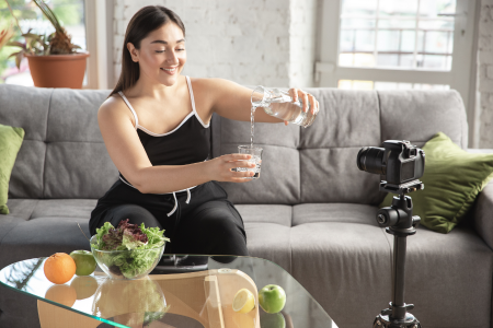 fit healthy latin woman wearing black sweatpants and black tank top sitting on her grey couch pouring water into a glass in front of her camera at her home