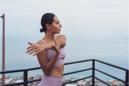 fit healthy latin woman wearing pink leggings and pink sports bra stretching outside on top of a balcony overlooking the ocean