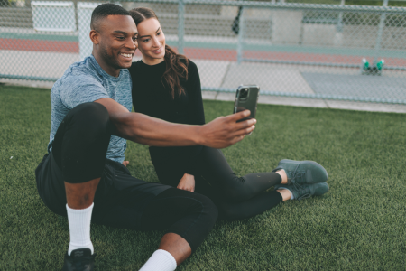 a black man wearing black jogging pants and grey t-shirt taking a pic with a white woman wearing all black activewear on the field of a stadium