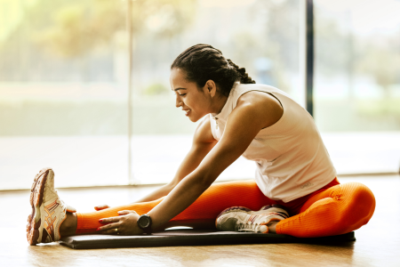 fit healthy latin woman wearing orange leggings and white tank top stretching her legs on her yoga mat at her home gym