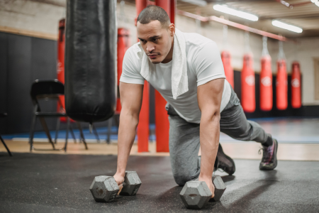 young man wearing a light grey t-shirt and grey pants with a towel around his neck doing push-ups with dumbbells on a floor at an MMA gym