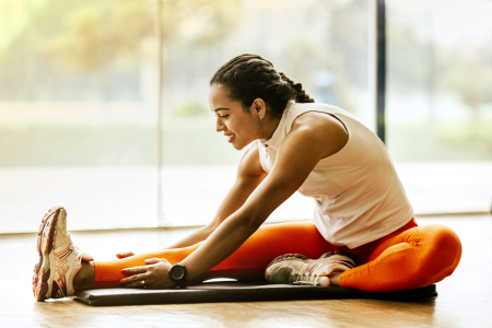 fit healthy latin woman wearing orange leggings and white tank top stretching her legs on a yoga mat at her home